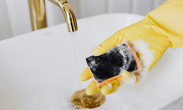 A photo of a person wearing a yellow glove holding a scrubbing pad covered in bubbles. Person is cleaning in hot soapy water.